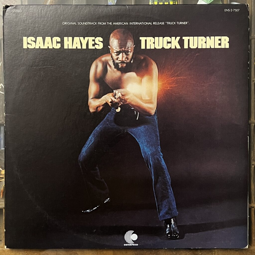 Truck Turner front cover – photograph of Isaac Turner, a tall, muscular Black man, bald headed, tight beard, facing the camera. he is standing in a bit of a crouch, legs apart and knees bent. He is wearing no shirt, dark blue jeans and shiny leather shoes. He is holding a firearm which is currently being fired, shown through a burst of bright flash at the muzzle. 