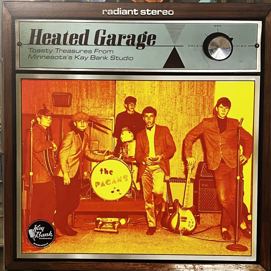 Heated Garage front cover – the top of the record is designed to look like a space heater, with the title on the left of the control panel and a temperature dial on the right, turned nearly to "high." Beneath that, there is a photograph of an all-male '60s garage band, The Pagans, five boys standing in a wood paneled hall with their instruments (guitars, drumset on a riser, amps and microphones).