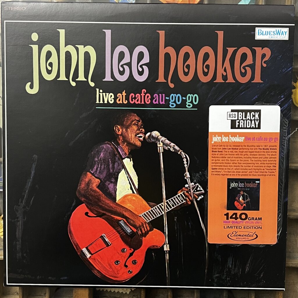 John Lee Hooker Live at Cafe Au-Go-Go front cover – the cover has john Lee Hooker's name in large, fantasy style serif letters; beneath that in much smaller letters is the album title, "live at cafe au-go-go." That is above an illustration of John Lee Hooker, standing in 3/4 profile at a microphone, singing into it, while playing a large f-hole electric guitar. The rest of the background is black.