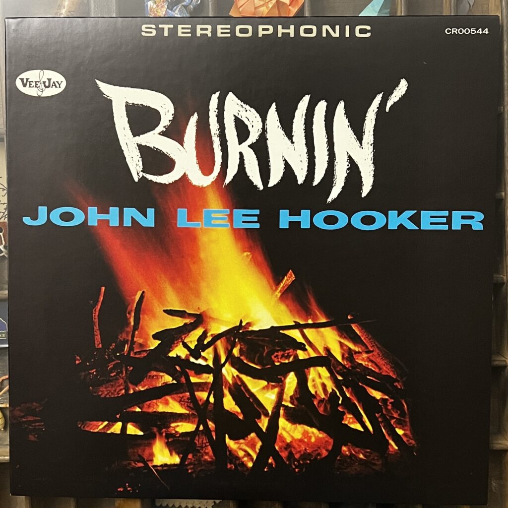 Front cover of John Lee Hooker's album "Burnin'" – at the top in a Eurostile font is the word "Stereophonic" and the catalog number, Below that, the Vee-Jay records logo, just the name in an oval with a G clef between Vee and Jay. Then in a white brush lettering, the title, Burnin'. Below that in a sans serif font, John Lee Hooker's name in blue. The whole thing is on a photograph of a wood fire, flames rising up through the title, mostly small pieces of wood, twigs and such, on fire below.