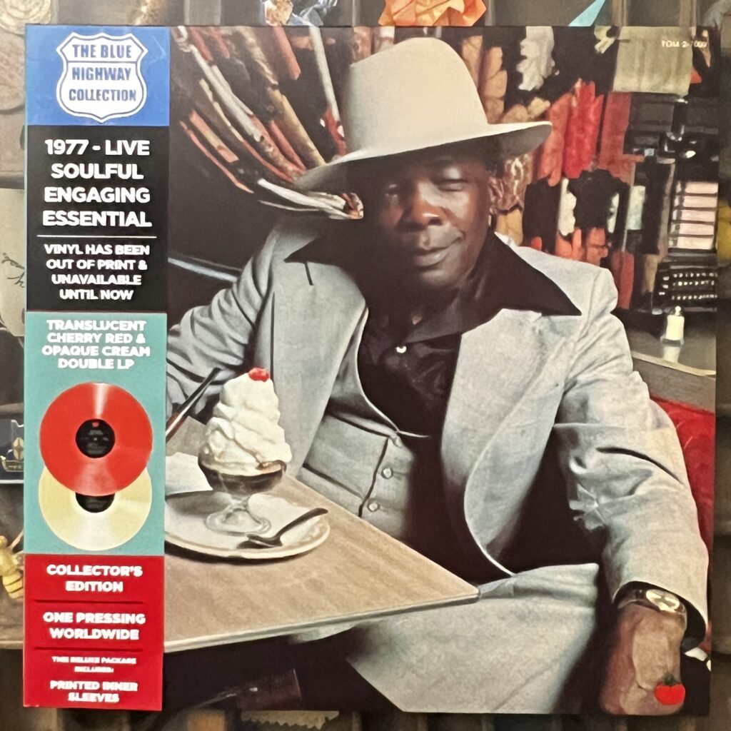 The Cream front cover – photograph of John Lee Hooker, a 60-something-year-old black man in a very dapper light colored three piece suit, a black silk or nylon shirt with an incredibly wide collar reaching to the shoulders of his suit jacket, and a tall, broad-brimmed light colored hat. He is seated at a diner table, a tabletop jukebox selector in the booth behind him. He has one arm resting on the table, the other on his knee, his left leg crossed on his right knee, he sits turned out from the booth. On the table is a heaping ice cream sundae covered in whipped cream with a cherry on top.