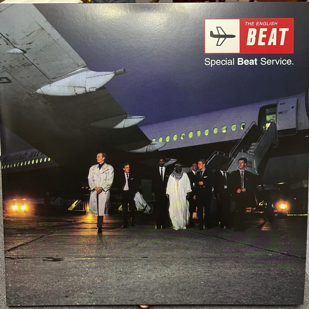 Cover of The English Beat, Special Beat Service. The cover is a photograph of the band in front of a jet airplane with an old style staircase to the tarmac. They appear to have just departed the plane, and are arranged roughly in a line, starting under the wing and across to the right. They are eight men, three Black and five white. The first is in a light colored raincoat; the rest are in dark suits with white shirts and narrow dark ties, except for a Black man in the middle who wears traditional Arab garb with a ghutra and perhaps (sorry, my cultural knowledge is limited) a thobe. The overall color cast is dusky blue.