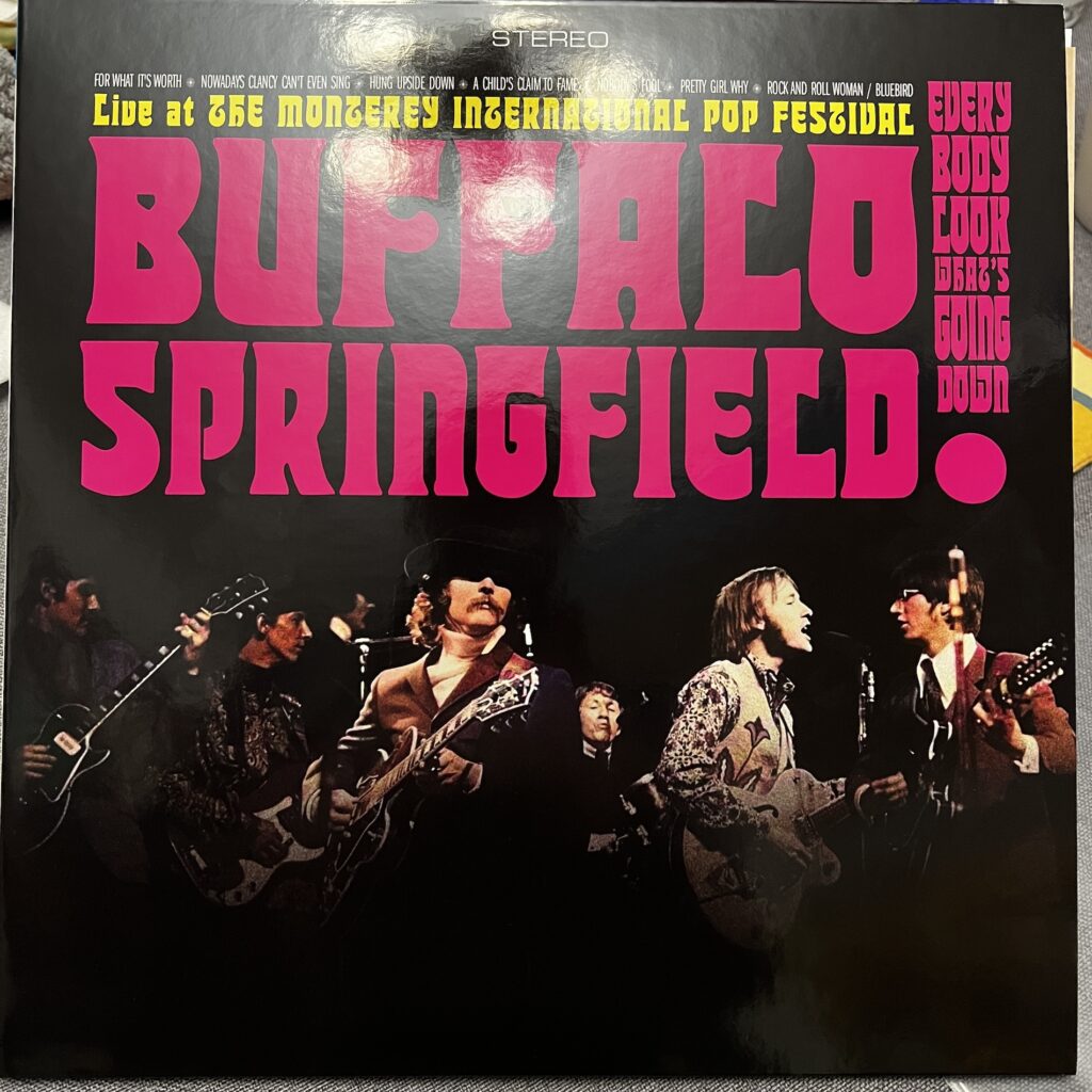 Cover of Buffalo Springfield at Monterey International Pop Festival, with the band's name in giant type covering the top half of the record, along with incidental type with track names, etc. Below that, a photograph of the band in the dark, six white men in fairly close proximity on a dark stage. To the left, in the shadow, a guitar (Neil Young) and bass player (Bruce Palmer). Near center and more prominent, a mustache-bearing guitar player, David Crosby, sitting in with the band for this performance. In the background, a drummer, Dewey Martin. to the right, and facing each other, Stephen Stills and Richie Furay on his twelve-string.  