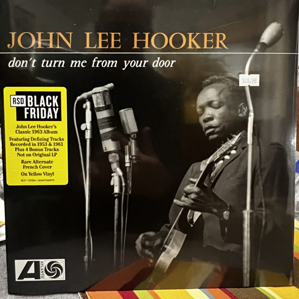 cover of John Lee Hooker, "Don't Turn Me From Your Door." The type is across the top. The cover is a black and white photograph of John Lee Hooker, standing, wearing a dark suit, playing guitar before a series of microphones. He is to the right side of the photo, facing the left, in a three-quarter turn, his eyes cast down toward his guitar but perhaps entirely closed as he plays. 