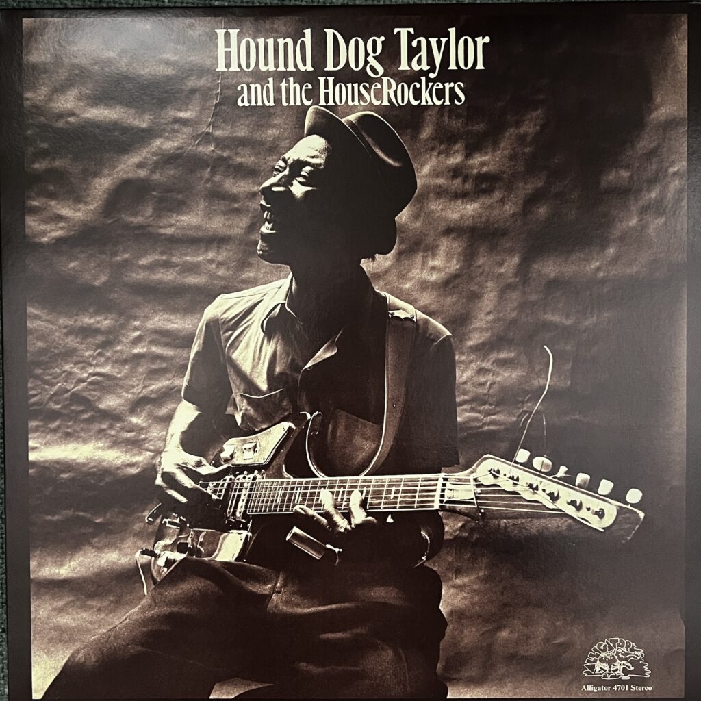 Hound Dog Taylor and the Houserockers front cover showing Hound Dog Taylor, a black man with a big smiling expression, wearing a jaunty narrow-brimmed hat, looking off to his right as he plays an electric guitar, seated, with a slide on his pinky finger. He is photographed in front of a rough backdrop. The photo is in black and white.