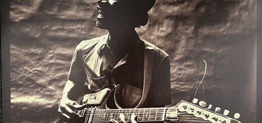 Hound Dog Taylor and the Houserockers front cover showing Hound Dog Taylor, a black man with a big smiling expression, wearing a jaunty narrow-brimmed hat, looking off to his right as he plays an electric guitar, seated, with a slide on his pinky finger. He is photographed in front of a rough backdrop. The photo is in black and white.