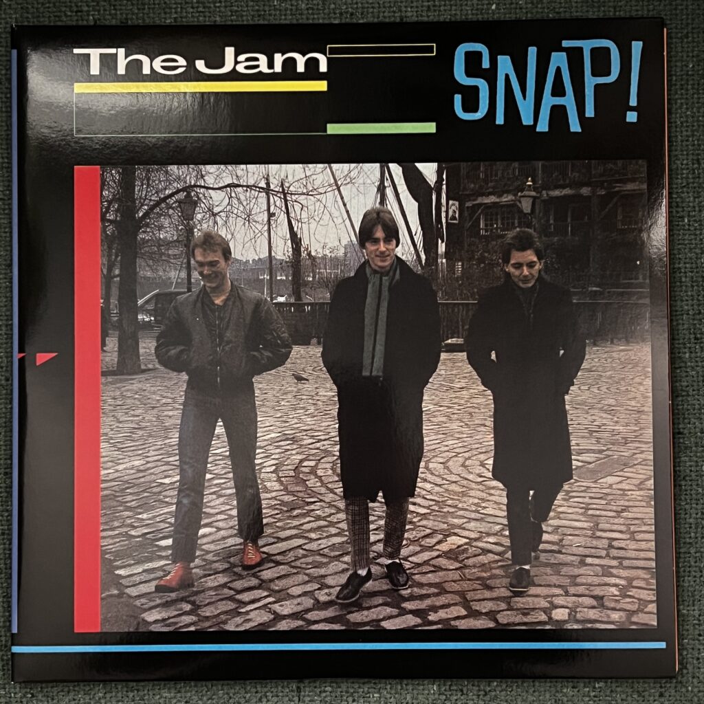 The Jam – Snap! front cover. The top of the album features the band name and title in jaunty font. Beneath that is a picture of three members of the Jam, three young men, walking toward the viewer in cold weather gear, all with hands in their pockets, crossing a gray public square of belgian pavers – one in a short jacket; the middle on in long black coat and gray scarf; the third also in long black coat. 