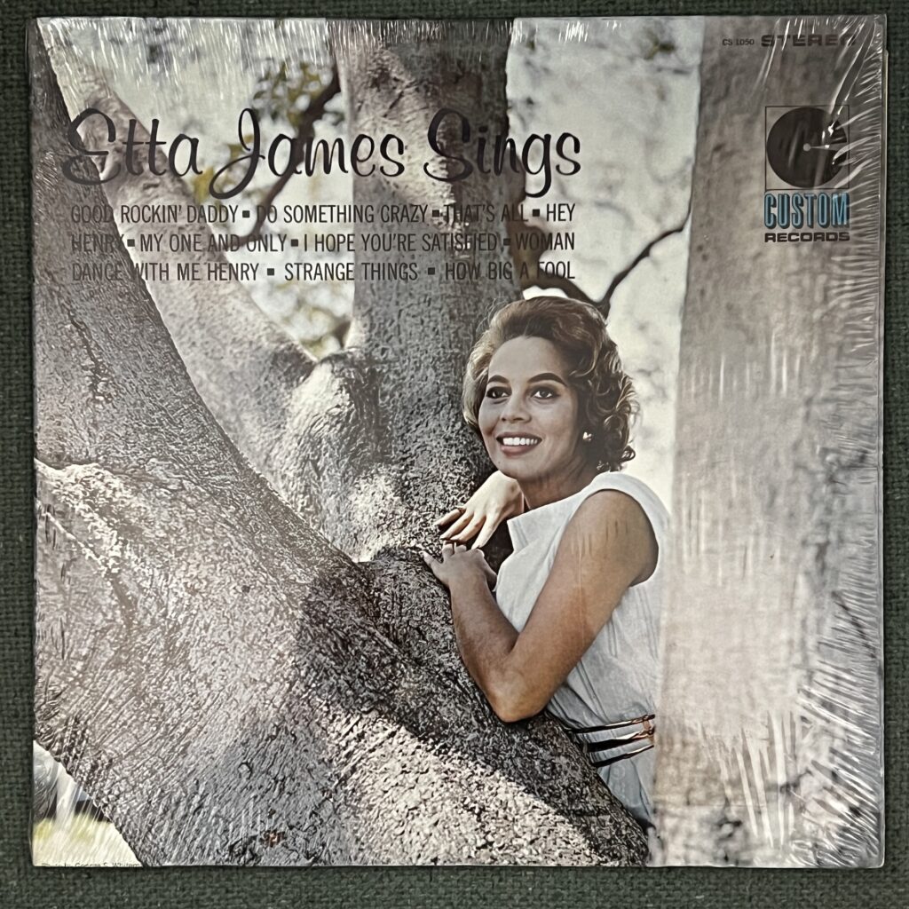 Etta James Sings front cover – photograph of Etta James, a black woman with light-colored wavy hair arranged back from her face in a loose '50s hairstyle, gazing into the distance with a bright smile. She is posed leaning against an enormous tree, her arms resting atop a spreading limb. She is wearing a white dress with light striping and a metal belt. 