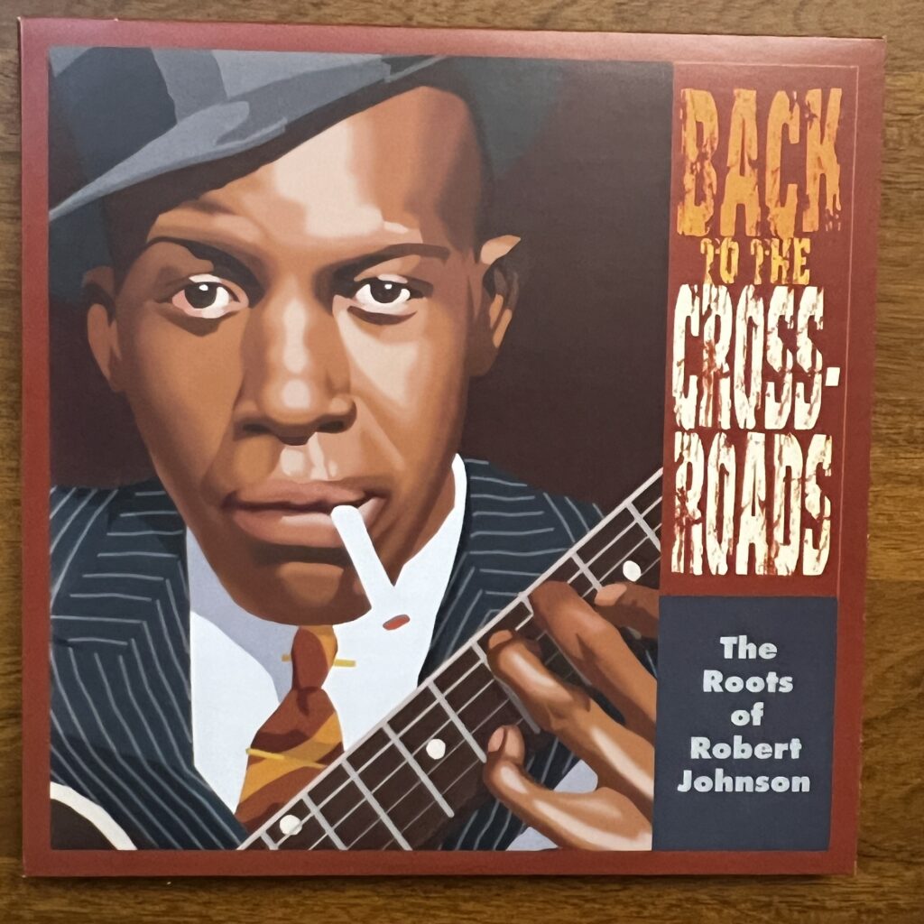 front cover of the album Back to the Crossroads, the Roots of Robert Johnson. The cover is dominated by a posterized version of a photograph of Robert Johnson, showing a Black man wearing a tilted fedora, staring directly at the viewer, cigarette dangling from his lip. He is wearing a sharp pin-striped suit jacket, white shirt with striped tie, and the neck of his guitar is just visible across the lower corner of the picture, his fingers just in evidence on the fretboard.