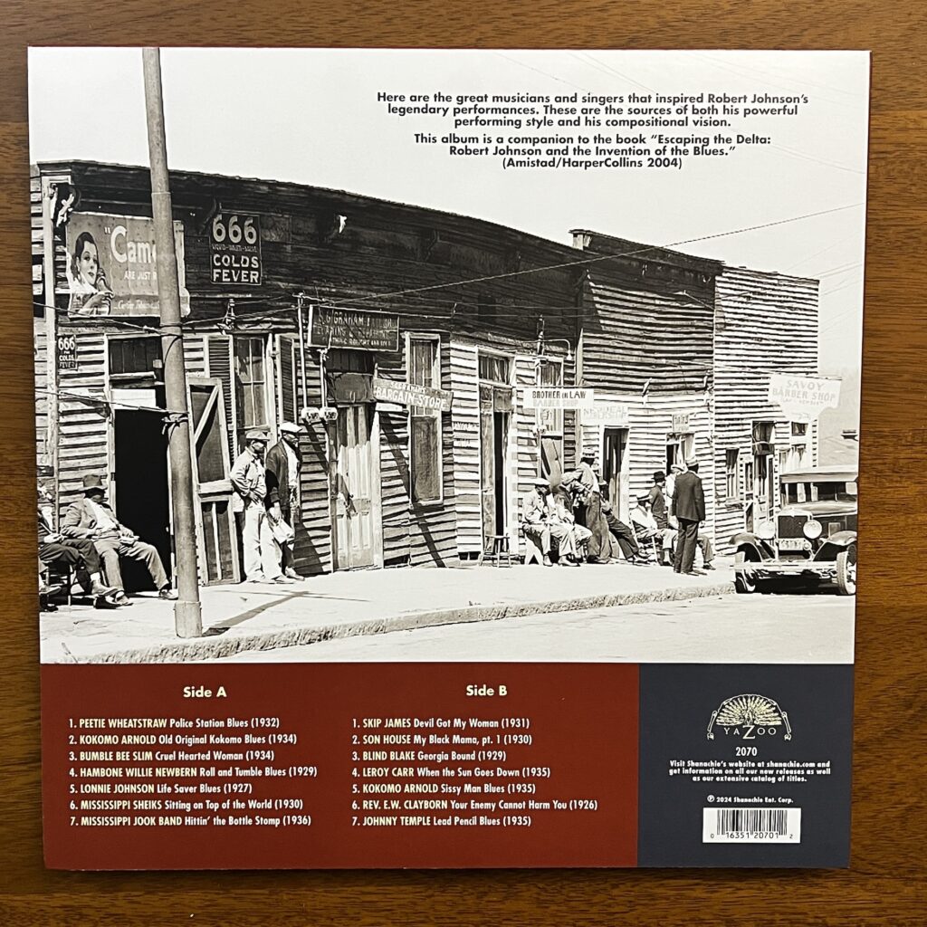 The back cover of Back to the Crossroads, featuring the track listing and a large photograph of a rather ramshackle set of commercial buildings, presumably taken in the 1930s. They are a row of wood frame buildings, all about eh same height, about a story and a half, sided in rough clapboard, and aged into odd angles. They include signage for a bargain store and two barber shops. A number of black men are standing or sitting outside of the shops.