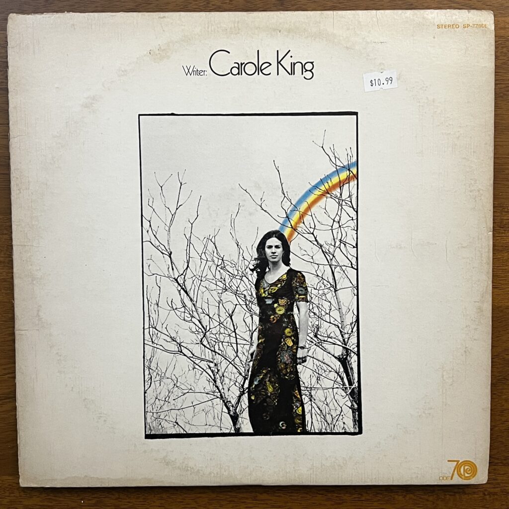 Front cover of Writer by Carole King. Her name is emphasized over the album title. Below that is a rectangular photograph of Carole King, standing in a long, floral dress, before the upper reaches of bare-limbed trees, a drawn three-color rainbow extending from the back of her head to the sky. The only color in the picture is in the dress and the rainbow.