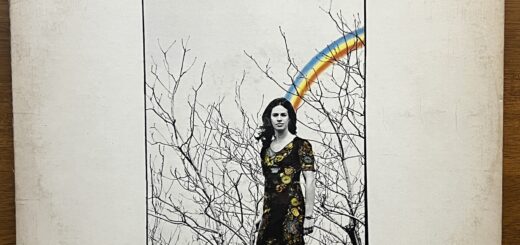 Front cover of Writer by Carole King. Her name is emphasized over the album title. Below that is a rectangular photograph of Carole King, standing in a long, floral dress, before the upper reaches of bare-limbed trees, a drawn three-color rainbow extending from the back of her head to the sky. The only color in the picture is in the dress and the rainbow.