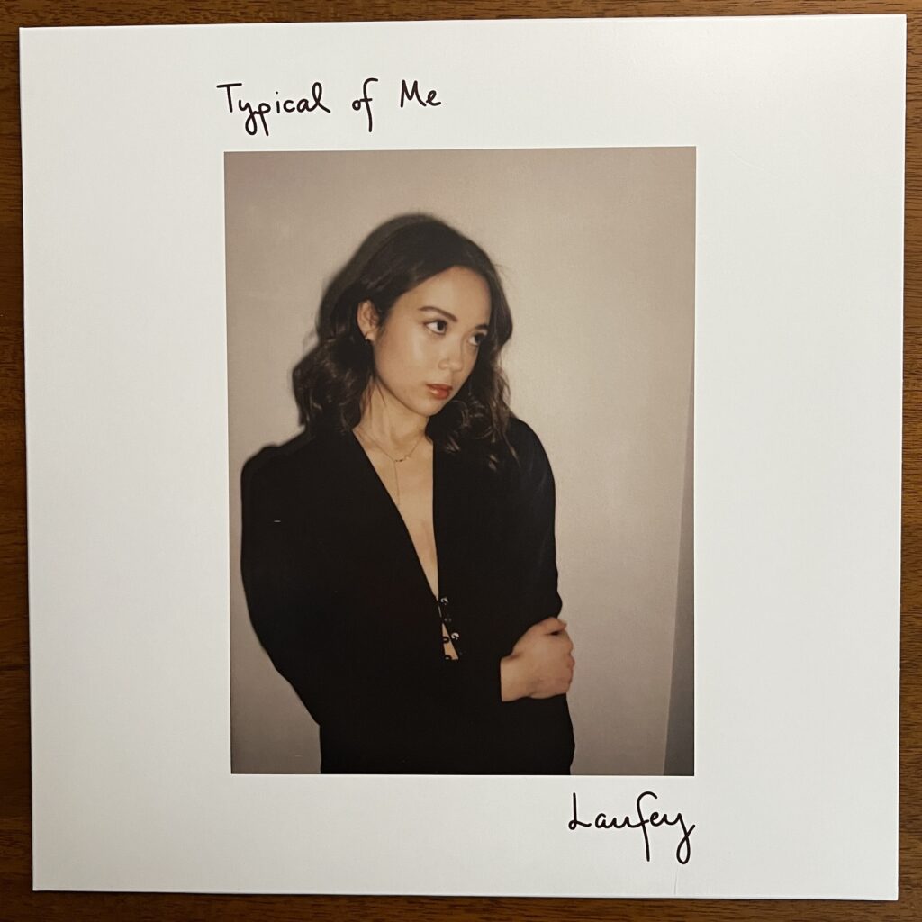 Typical of Me front cover – the title and name of the artist, Laufey, are written in a simple pen text. Between them is a photograph of the singer, appearing to be a polaroid, with rather flat colors. She is posed against a blank wall, harsh shadow behind her. She is looking to her left, her head tilted down toward her left shoulder; her right arm is lightly clutching her left elbow. She is wearing a dark, low-cut top that is held closed only by two frogs about midway down. Two more frogs are open, exposing more skin below.