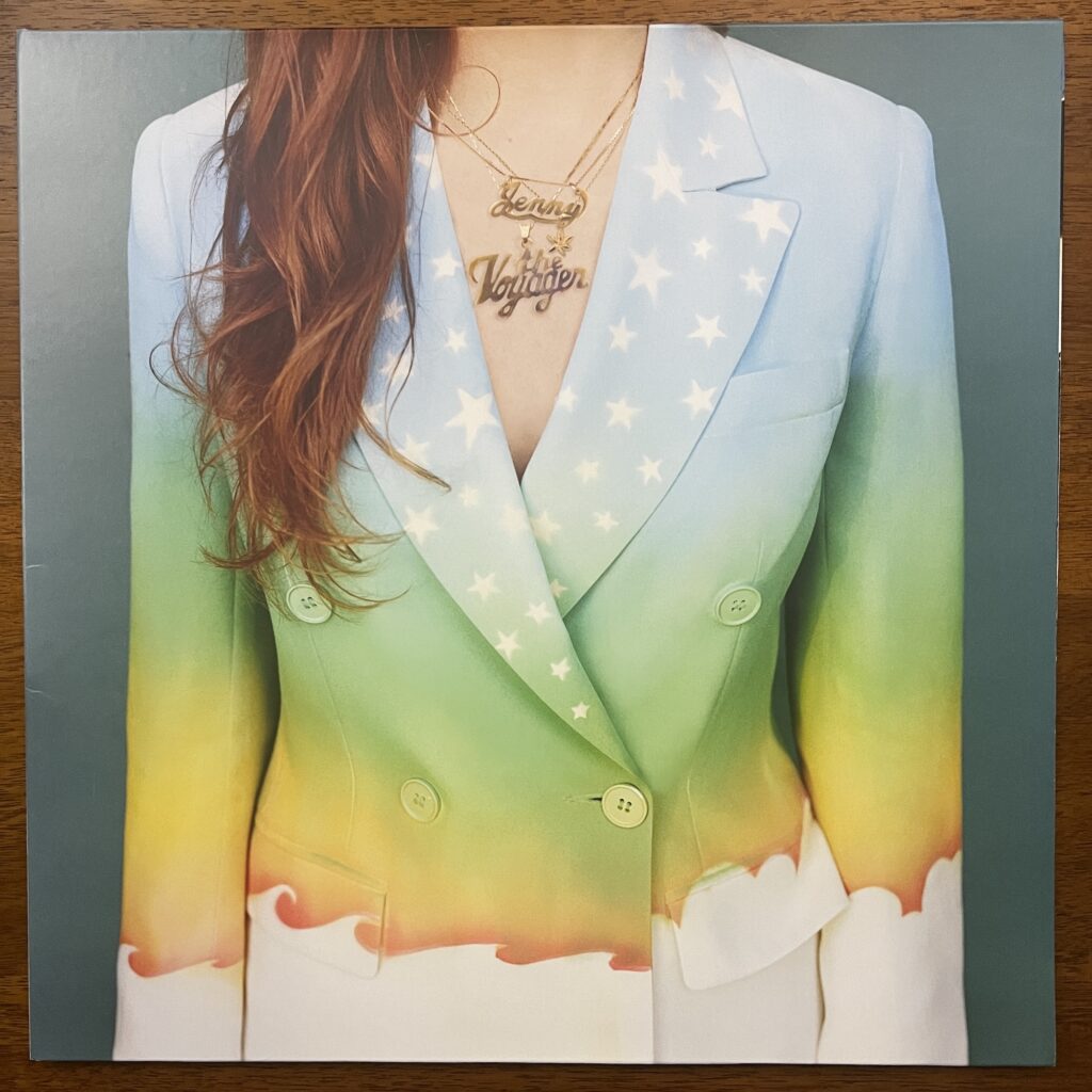 Jenny Lewis Voyager front cover, showing just a woman's torso in a colorful blazer, perhaps painted. It is light colored at the top, blue shifting to darker green moving down the jacket, then turning to white with a line of ocean waves and clouds. There are stars across both lapels. Her brown hair hangs down on her right side, lightly wavy. Her bare skin shows between the lapels, with a necklace that is cut-out letters reading "Jenny" above and "The Voyager," the album title, below.