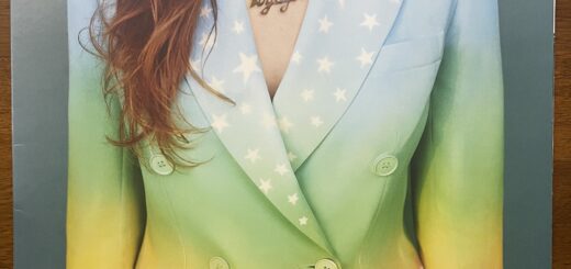 Jenny Lewis Voyager front cover, showing just a woman's torso in a colorful blazer, perhaps painted. It is light colored at the top, blue shifting to darker green moving down the jacket, then turning to white with a line of ocean waves and clouds. There are stars across both lapels. Her brown hair hangs down on her right side, lightly wavy. Her bare skin shows between the lapels, with a necklace that is cut-out letters reading "Jenny" above and "The Voyager," the album title, below.