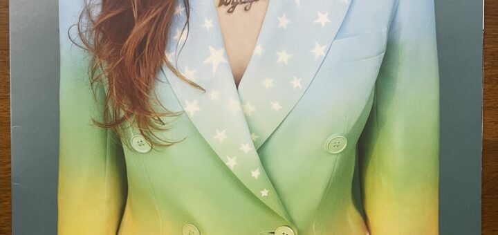 Jenny Lewis Voyager front cover, showing just a woman's torso in a colorful blazer, perhaps painted. It is light colored at the top, blue shifting to darker green moving down the jacket, then turning to white with a line of ocean waves and clouds. There are stars across both lapels. Her brown hair hangs down on her right side, lightly wavy. Her bare skin shows between the lapels, with a necklace that is cut-out letters reading "Jenny" above and "The Voyager," the album title, below.