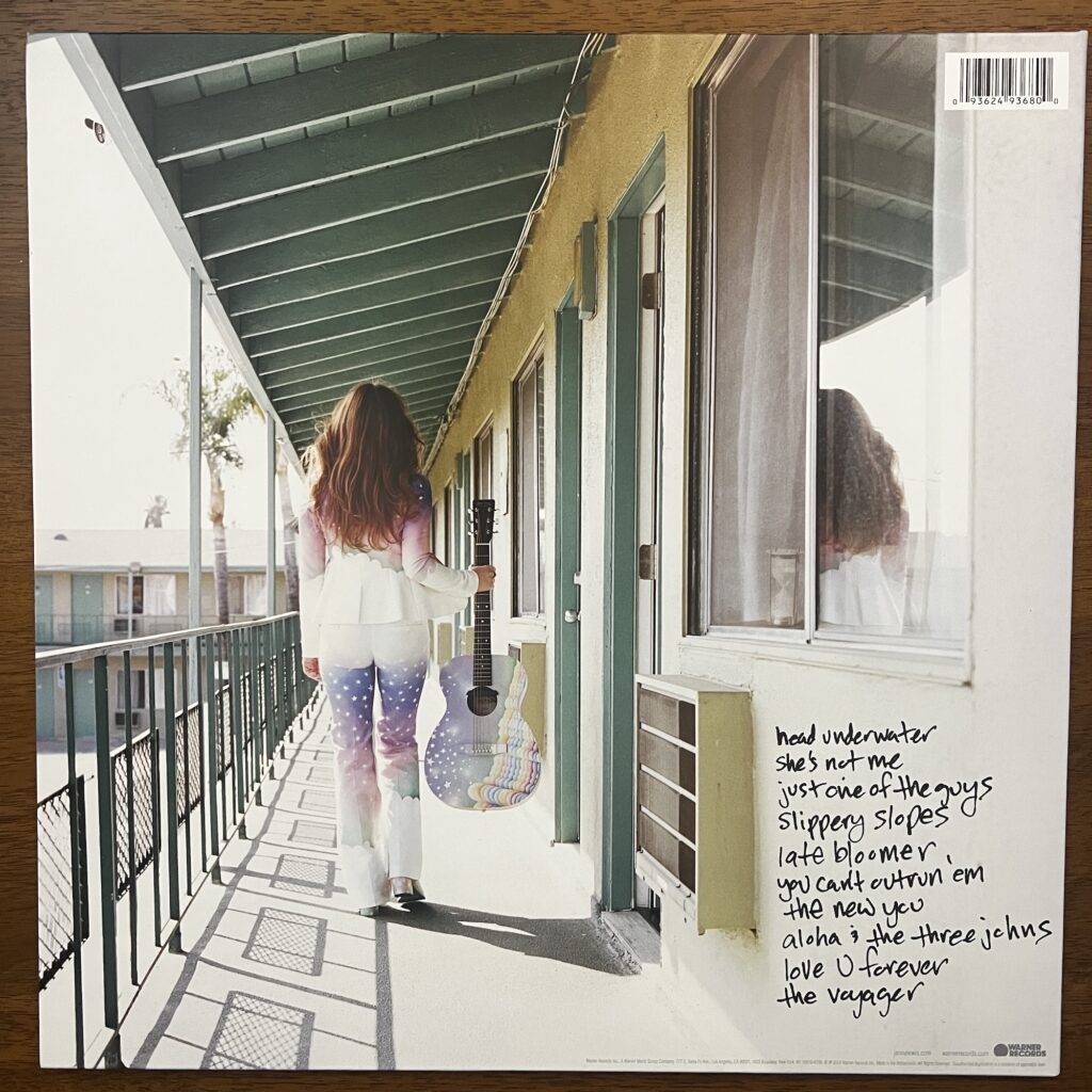 Voyager back cover, showing Jenny Lewis from behind, walking away from us on the upper deck of a motel, passing the row of doors and windows, steel railing on her left. She is wearing a colorful outfit, with a blazer on top and pants below that are painted with stars and sky imagery. She is carrying an acoustic guitar, front facing us, in her right hand, and it is painted like her clothes, with stars and sea/sky colors.