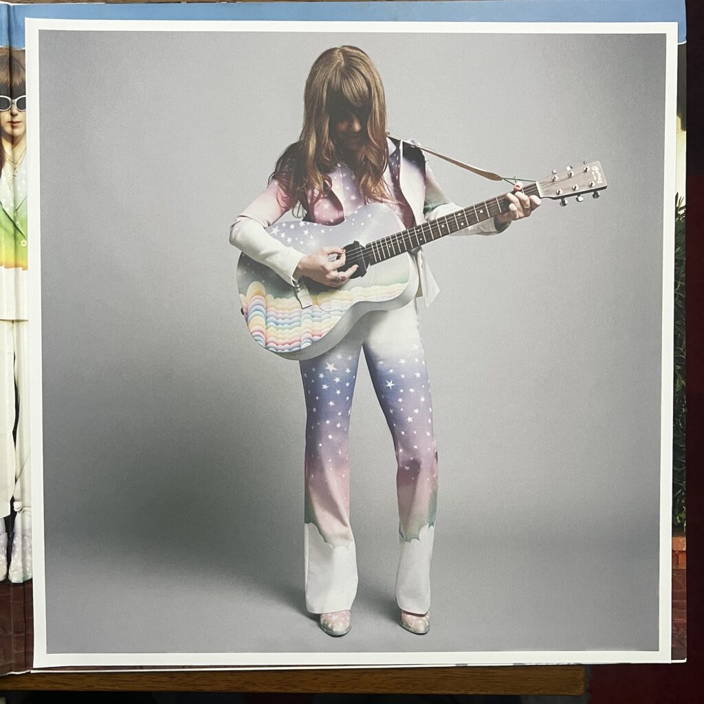 Voyager insert, showing Jenny Lewis playing her acoustic guitar. Both her clothes and the guitar are painted with light sky and sea colors, and decorated with stars, waves and clouds.