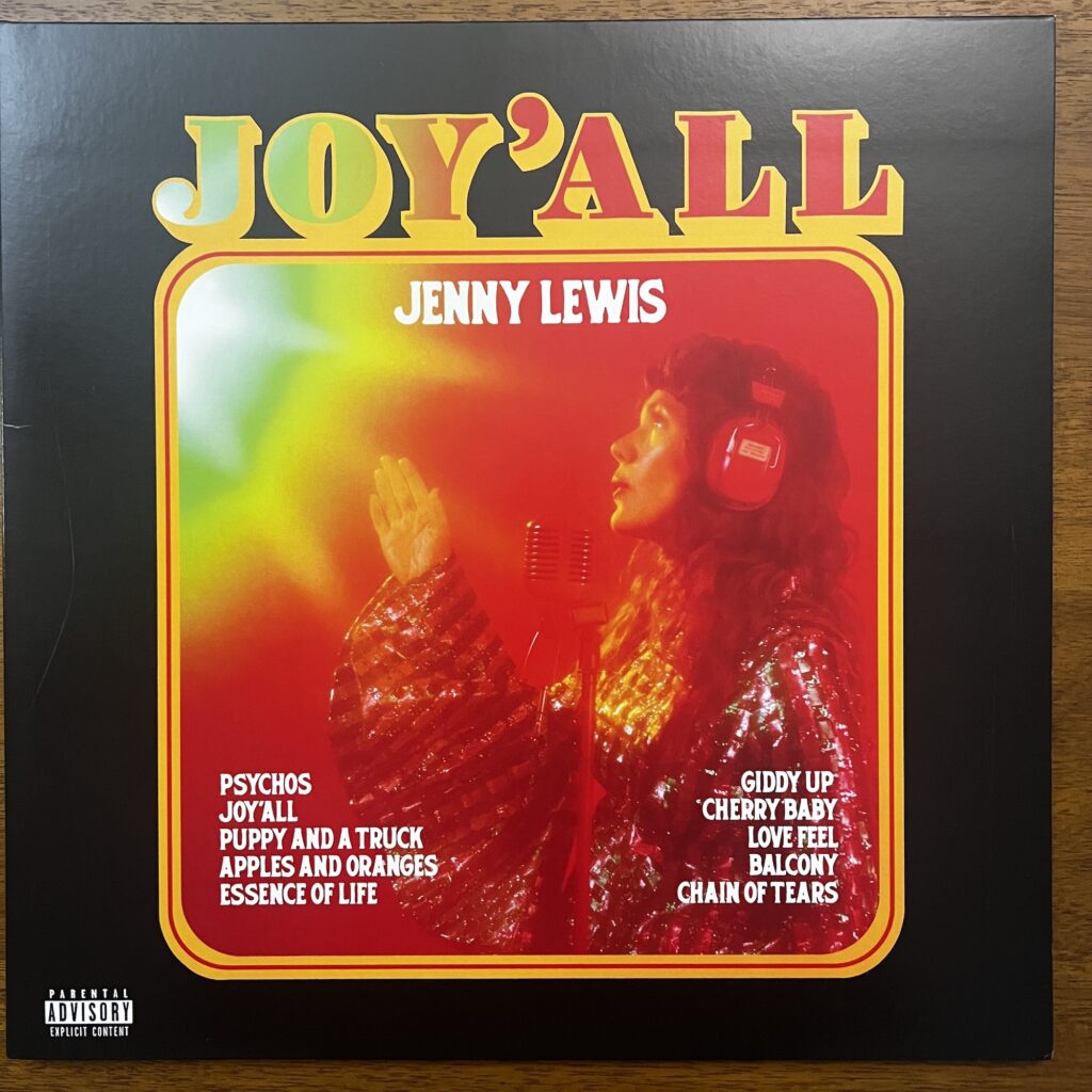 Joy'All front cover. The title is in large profiled type at the top, beneath which is a round-cornered near square with a photograph of Jenny Lewis in profile, wearing headphones and singing into a studio microphone, wearing a very glittery top. The song titles are listed in the corners of the photo box.