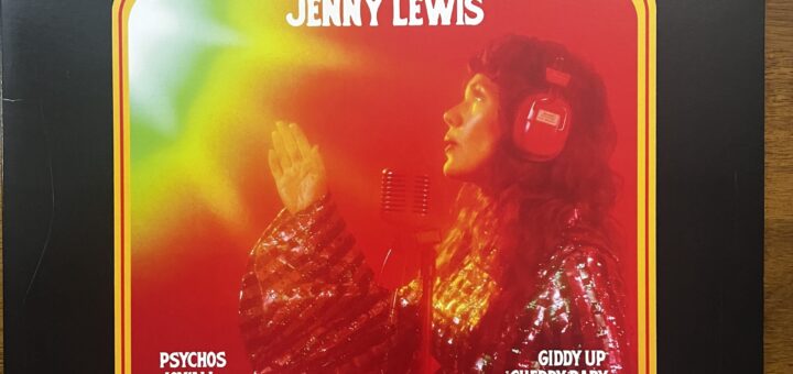 Joy'All front cover. The title is in large profiled type at the top, beneath which is a round-cornered near square with a photograph of Jenny Lewis in profile, wearing headphones and singing into a studio microphone, wearing a very glittery top. The song titles are listed in the corners of the photo box.