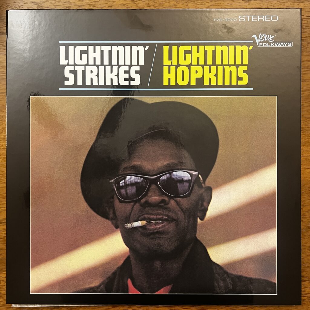 Lightnin' Strikes front cover – featuring the title stacked to the left, and then Lightnin' Hopkins stacked to the right. Below that is a picture of Hopkins, a middle-aged Black man, wearing a cocked hat, sunglasses, and smoking a cigarette, looking straight at the viewer.