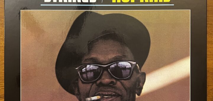 Lightnin' Strikes front cover – featuring the title stacked to the left, and then Lightnin' Hopkins stacked to the right. Below that is a picture of Hopkins, a middle-aged Black man, wearing a cocked hat, sunglasses, and smoking a cigarette, looking straight at the viewer.