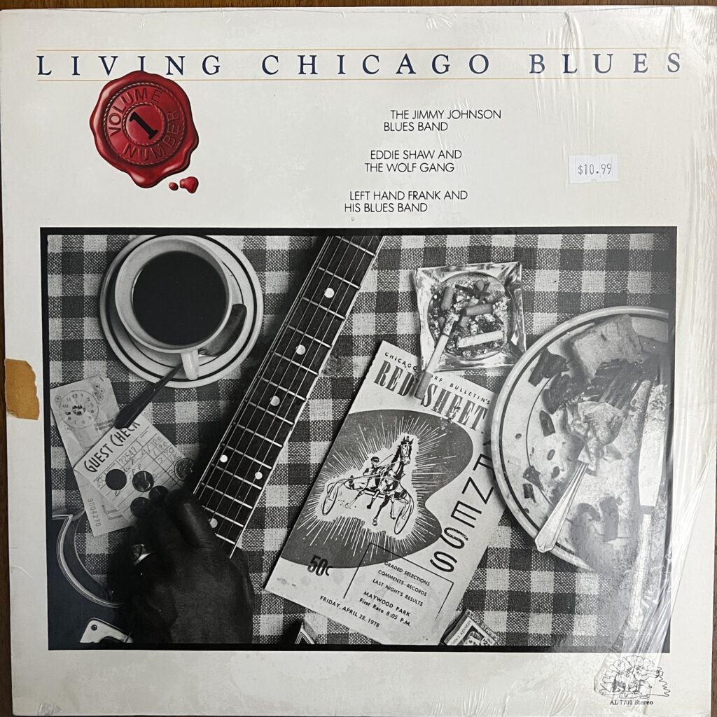 Living Chicago Blues Vol. 1 – The album cover features a banner across the top reading "Living Chicago Blues" in a nice Goudy font, underneath which is a graphic of a red wax seal that reads "volume number 1." The names of the bands featured are to the right of the seal in a modest sized sans serif font – The Jimmy Johnson Blues Band, Eddie Shaw and the Wolf Gang, and Left Hand Frank and His Blues Band. Beneath that is a photograph of what appears to be a restaurant table setting, with a checkered tablecloth, atop which sits a cups of coffee with spoon, an ashtray full of spent cigarettes, a plate  with fork and knife and some food remnants, a "red sheet" horse racing publication. Across the left side is the neck of an electric guitar, held by an unidentified black hand wearing a gold ring on his ring finger.