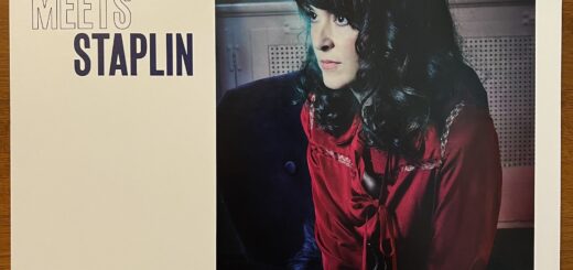 April March Meets Staplin front cover – title type is on the left side, with a rectangular photograph of April March to the right. She appears perched on the edge of an upholstered chair, looking off to the left of the frame in 3/4 profile. She has dark, curled long hair falling below her shoulders, white skin and dark, round eyes. She is wearing a red top with decorative trim forming a yoke, a narrow band of fabric forming a loose bow, and a studded belt. behind her is an old-style perforated radiator cover and patterned curtains over a window, partially seen.
