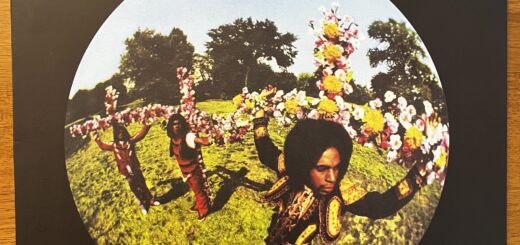 Maximillian front cover, featuring a round, fish-eye lens view of the three band members, wearing very colorful regalia and positioned on crucifixes covered in flowers.