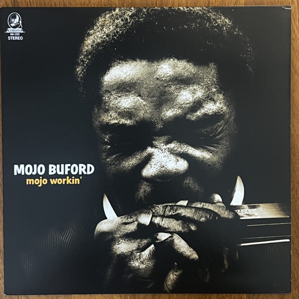 The cover of Mojo Buford's "mojo workin'" album , with the title to the left below center. The photograph is a head-on shot of Mojo Buford, a black man with short natural hair, his eyes squinted shut as he blows into a harmonica held tight in his hands.