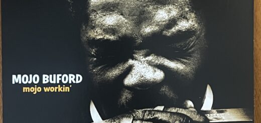 The cover of Mojo Buford's "mojo workin'" album , with the title to the left below center. The photograph is a head-on shot of Mojo Buford, a black man with short natural hair, his eyes squinted shut as he blows into a harmonica held tight in his hands.