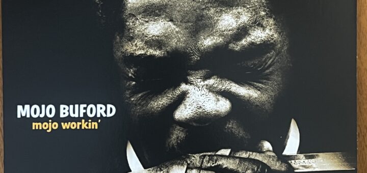 The cover of Mojo Buford's "mojo workin'" album , with the title to the left below center. The photograph is a head-on shot of Mojo Buford, a black man with short natural hair, his eyes squinted shut as he blows into a harmonica held tight in his hands.