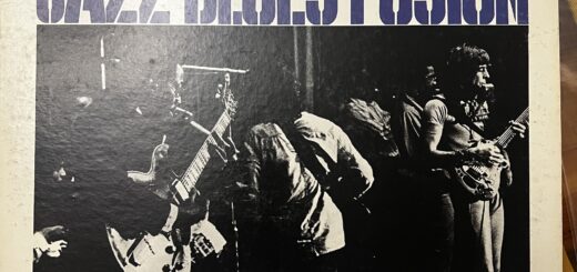 The cover of John Mayall's Jazz Blues Fusion album. The title above the photo, and more text below that says "performed and recorded live in Boston and New York," are rendered in a stencil font. The photograph is rectangular black and white, showing Mayalll's band playing, showing a black man (Freddy Robinson) playing electric guitar, anodht erplayer with his back to us, and John Mayall at the far right, barely in the picture, playing guitar and with a harmonica in a holder near his mouth. Other players are barely visible in the background.